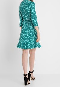 Tyrkysové šaty s bílými puntíky, s 3/4 rukávy, se zúženým pasem, rozšířeným lemem a klíčovou dírou na zádech. V kombinaci s černými sandály na blokovém podpatku.