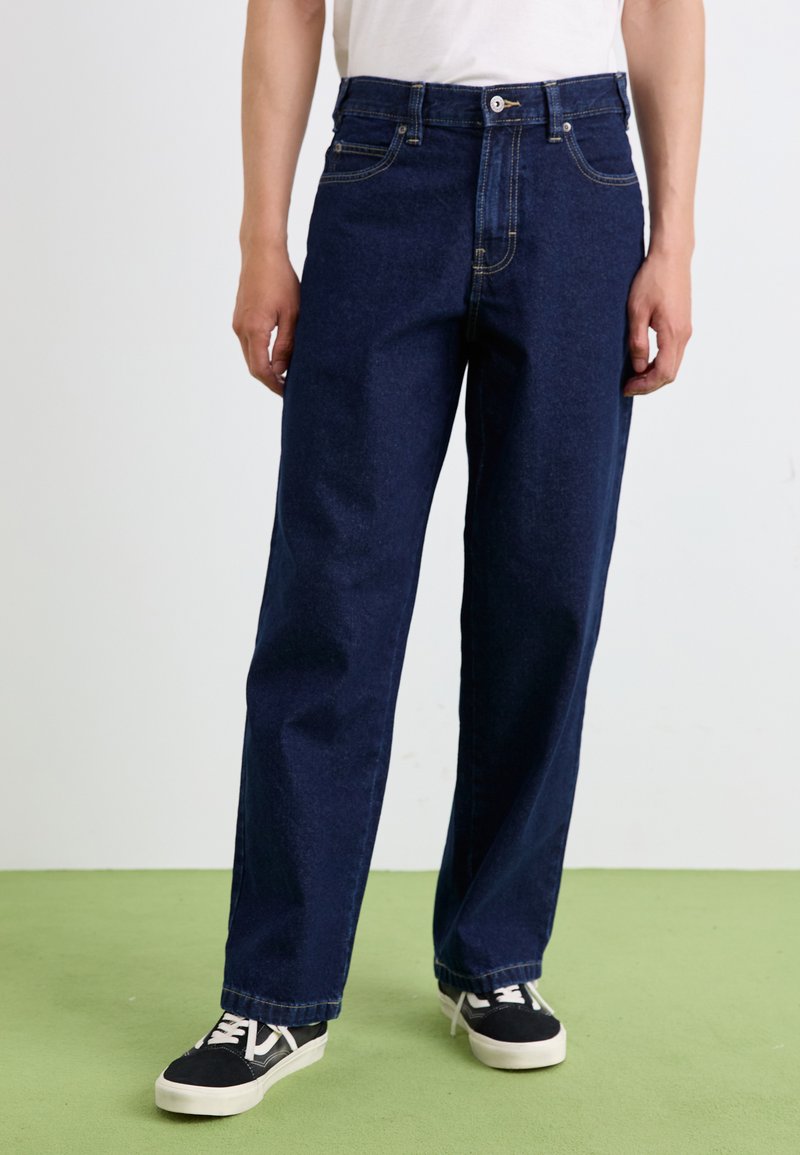 Jeans en denim bleu foncé avec une coupe droite, dotés d'une fermeture à bouton, de cinq poches et de détails de couture subtils. Accompagnés de baskets noires.