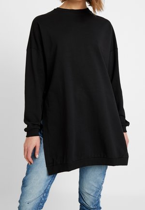 Felpa nera oversize in cotone morbido con maniche lunghe, scollo rotondo e spacchi laterali, abbinata a jeans in denim azzurro chiaro.