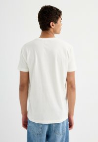 Camiseta de algodón blanca con cuello redondo y mangas cortas, que presenta un corte holgado. La vista trasera muestra una tela lisa sin gráficos ni patrones.