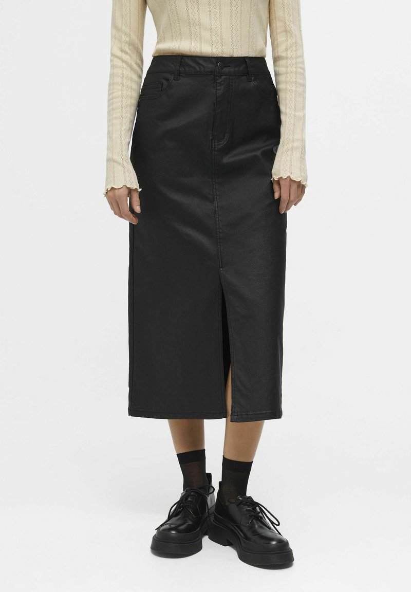 Jupe crayon noire longueur genou en simili cuir avec une fente à l'avant, associée à des chaussures à plateforme noires et un haut en tricot beige clair.