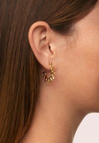 Orecchini a cerchio dorati con accenti a forma di foglia, caratterizzati da una superficie texture e un design irregolare. Posizionati sull'orecchio di un modello.