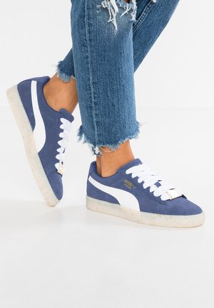 Pieds chaussés de baskets Puma en daim bleu avec des lacets blancs et des bandes blanches, associés à un jean bleu à ourlet effiloché sur un fond blanc.