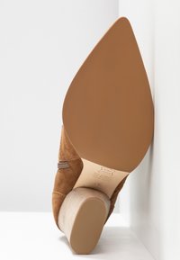 Chaussure à talon haut en daim marron avec un talon texturé beige et une semelle lisse. Elle dispose d'une fermeture éclair sur le côté et de détails en surpiqûre sur le dessus.