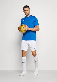 Vyras mėlyna marškinėliais, baltais šortais ir kojinėmis, laikantis auksinį futbolo kamuolį, stovintis priešais paprastą baltą foną.