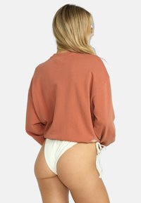 Rustfarget oversized hettegenser med lange ermer, som har en løs passform og justerbar knyting i bunnen. Hvit teksturert bikinibunn.