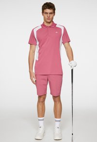 Rosa polotröja med vita sidopaneler, matchad med rosa shorts. Har en krage, logotyp på bröstet, och modellen håller en golfklubba, iklädd handskar.