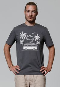T-shirt en coton gris avec un imprimé graphique blanc d'une van, de palmiers, et le texte « Que l'aventure commence ! » sur un fond neutre.