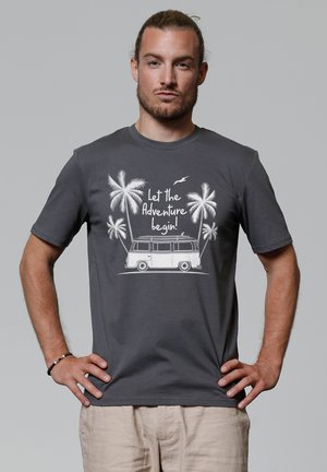 Grijze katoenen t-shirt met witte grafische print van een bus, palmbomen en de tekst "Laat het avontuur beginnen!" op een neutrale achtergrond.