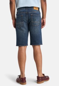 Pantalones cortos de mezclilla en un tono azul oscuro, con un corte recto, bolsillos traseros y una etiqueta de cuero marrón en la cinturilla. El calzado son mocasines de color rojo oscuro.