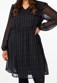 Svart klänning med genomskinliga långa ärmar, V-ringning och volangdetaljer. Har horisontella vita ränder och en samlad elastisk midja.