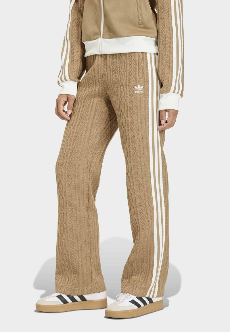Calças tricotadas em castanho com riscas laterais brancas e o logótipo da adidas. Combinadas com ténis brancos com riscas pretas.