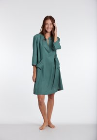 Teal groene jurk van een gladde stof, met een diepe V-hals, driekwartmouwen en twee zijzakken, in een relaxte pasvorm.