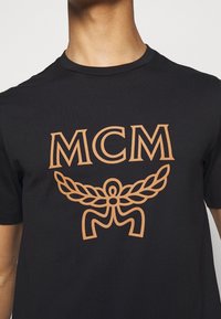 Camiseta de algodón negra con un gran logo de MCM en color beige, decorado con un emblema de hojas y un motivo central. Diseño de manga corta y cuello redondo.
