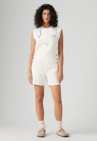 Salopette en denim blanche avec des bretelles réglables et des boutons sur les côtés, associée à un débardeur blanc et des chaussettes couleur crème.