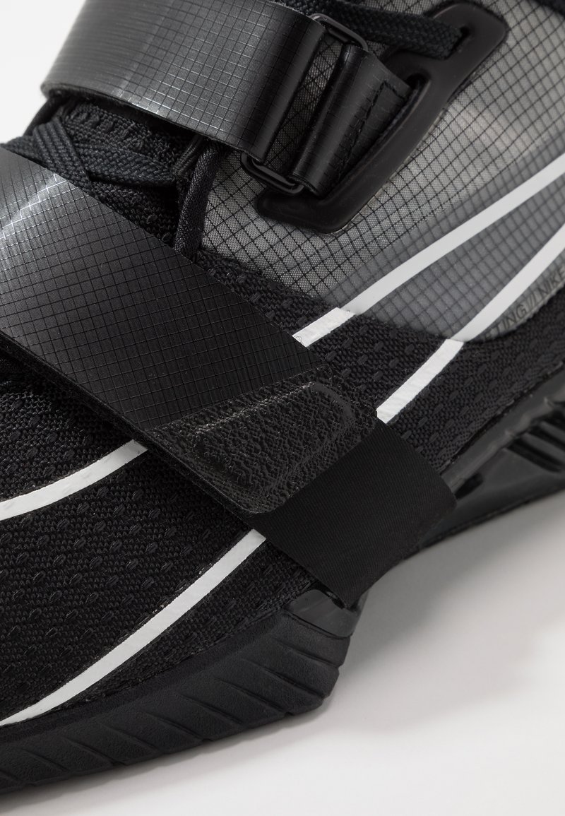 Czarne buty sportowe z teksturowaną cholewką i siateczkowymi panelami, białymi akcentami oraz regulowanymi paskami zapewniającymi pewne dopasowanie i wsparcie.