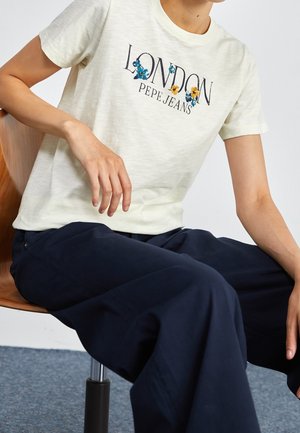 Lichtgeel T-shirt met korte mouwen met "LONDON" en bloemenborduursel; gecombineerd met marineblauwe broek met brede pijpen. Houten stoel met metalen onderstel.