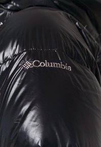 Veste matelassée noire avec une texture brillante, portant le logo Columbia brodé en argent sur la poitrine.