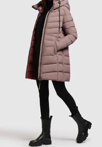 Femme portant une longue doudoune mauve avec capuche, des leggings noirs et des bottes noires à la cheville, se tenant avec une jambe en avant.