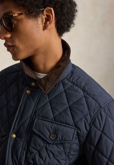 Jeune homme portant des lunettes de soleil en écaille de tortue et une veste matelassée bleu marine avec un col en velours marron, tourné vers la gauche sur un fond neutre.