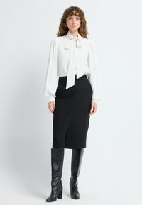Blouse blanche avec un col à nœud, manches volumineuses, jupe crayon noire et bottes noires texturées à mi-mollet. Design épuré et minimal.