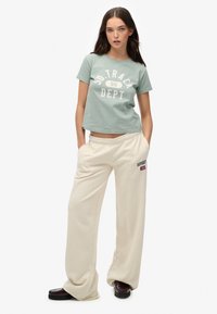 Lichtgroene cropped t-shirt met witte tekst "SD. TRACK DEPT 86." Crème-kleurige wijde sweatpants met logo op de zak.