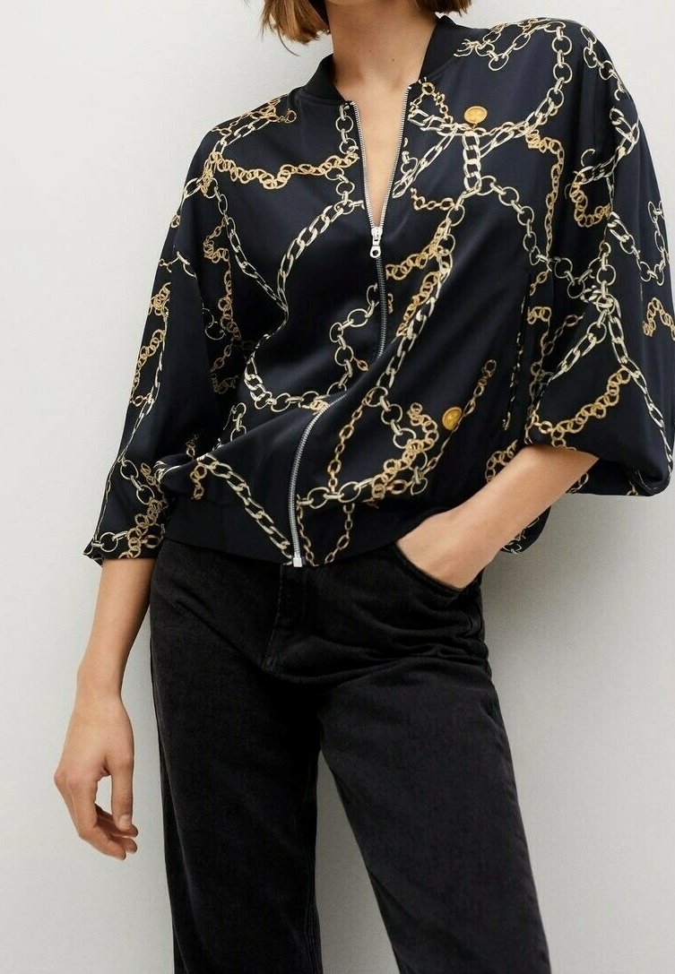 Chaqueta de satén negra con cremallera que presenta un patrón de cadenas doradas. Las mangas son anchas y cortas, con una textura suave y un brillo sutil.
