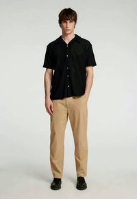Μαύρο πουκάμισο κοντό μανίκι με κουμπιά και υφή, συνδυασμένο με μπεζ παντελόνι και μαύρα παπούτσια, στέκεται μπροστά από απλό φόντο.