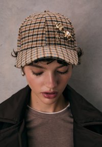 Cappello a quadri marrone con visiera curva, decorato con spille floreali dorate e dettagli blu. Tessuto morbido con finitura strutturata.