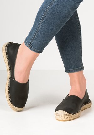 Fødder iført sorte espadrilles med flettede jutesåler, kombineret med cropped blå denimjeans, stående på en hvid overflade.