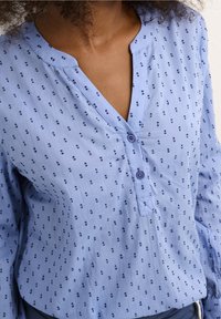 Blouse boutonnée bleu clair avec un col en V, présentant un motif discret de pois, manches longues et boutons d'un bleu légèrement plus foncé.