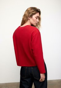 Maglioncino rosso corto con scollo rotondo e bordi a coste, caratterizzato da maniche lunghe e una trama liscia, abbinato a jeans scuri.