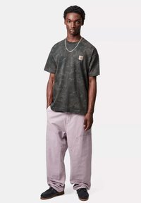 Ciemnozielona koszulka w kamuflażu z krótkim rękawem i naszywką, w połączeniu z luźnymi, jasnofioletowymi spodniami i czarnymi butami.