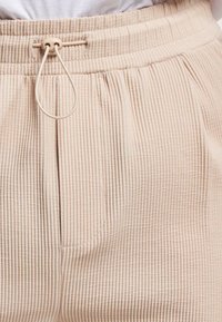 Pantalones de tela acanalada de color beige con cintura ajustable, que presentan plisados frontales y un bolsillo lateral, textura suave y diseño a medida.
