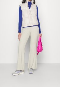 Beige padded vest over a blue turtleneck, cream wide-leg ribbed pants, gray sneakers with purple accents, holding a pink bag.