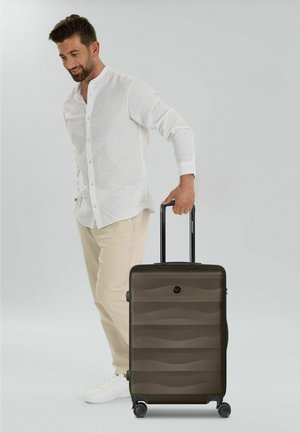 Uomo con camicia bianca e pantaloni beige che trascina una valigia rigida grigio scuro con quattro ruote su uno sfondo semplice.