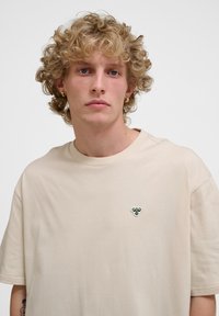 Beige bomuld t-shirt med en afslappet pasform. Har et lille dekorativt grønt og hvidt logo på venstre bryst. Korte ærmer og rund halsudskæring.
