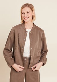 Giacca bomber marrone, leggera, con una texture liscia, bottoni a pressione e due tasche frontali. Indossata sopra una maglia bianca, leggermente luccicante.