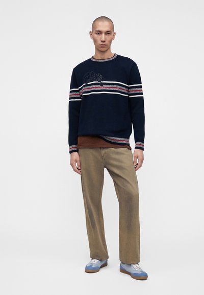 Homme portant un pull bleu marine à rayures rouges et blanches, pantalon beige, baskets bleues et blanches, debout devant un fond blanc uni.
