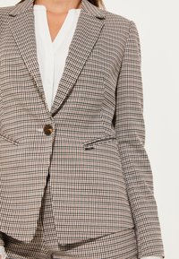 Blazer con motivo a quadri nei toni del marrone, beige e nero. Dettagli notevoli includono un bottone singolo, rever a scollatura e due tasche frontali.