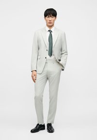 Άνδρας που φοράει ανοιχτό γκρι κοστούμι, λευκό πουκάμισο, σκούρα πράσινη γραβάτα και μαύρα παπούτσια, στέκεται με το ένα χέρι στην τσέπη.