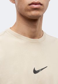 Κοντινό πλάνο στο κάτω μέρος προσώπου και λαιμό ενός ατόμου που φοράει μπεζ μπλούζα με μικρό μαύρο λογότυπο Nike swoosh.