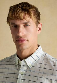 Weißes Hemd mit blauem und schwarzem Karomuster, Kentkragen und kurzen Ärmeln. Glatter Stoff mit leichtem Glanz, minimalistisches Design.