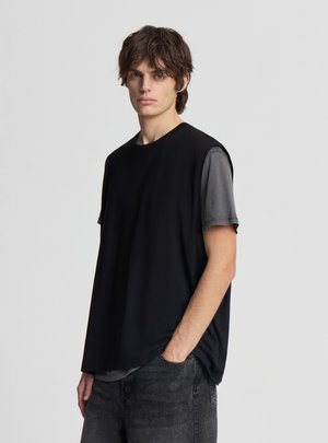 Młody mężczyzna z potarganymi brązowymi włosami, ubrany w czarny bezrękawnik na szarym krótkim rękawie i ciemne dżinsy, stojący na jednolitym tle.