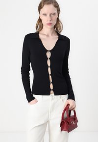 Zwarte lange mouw top met een V-hals en knoopdetails. Stof is glad; heeft een kraag en uitsnijding. Model houdt een rode handtas vast.