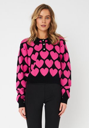 Roze hartpatronen op een zwart gebreide trui. Heeft een kraag met drie decoratieve knopen en lange mouwen. Getailleerde pasvorm.