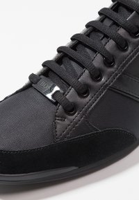 Czarny sneaker wyróżniający się połączeniem teksturowanej tkaniny i zamszu, z metalowymi oczkami, czarnymi sznurowadłami oraz markowym metalowym tagiem na języku.