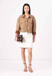 Bruine suède cropped jack met twee voorzakken, gelaagd over een rood geruit shirt, gecombineerd met een witte gewatteerde rok en een bruine tas.