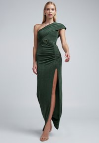 Jednokrčné zelené šaty z hodvábneho velúru so širokým rázporkom na boku, zdobené záhybmi a priliehavým strihom, doplnené dekoratívnym opaskom.