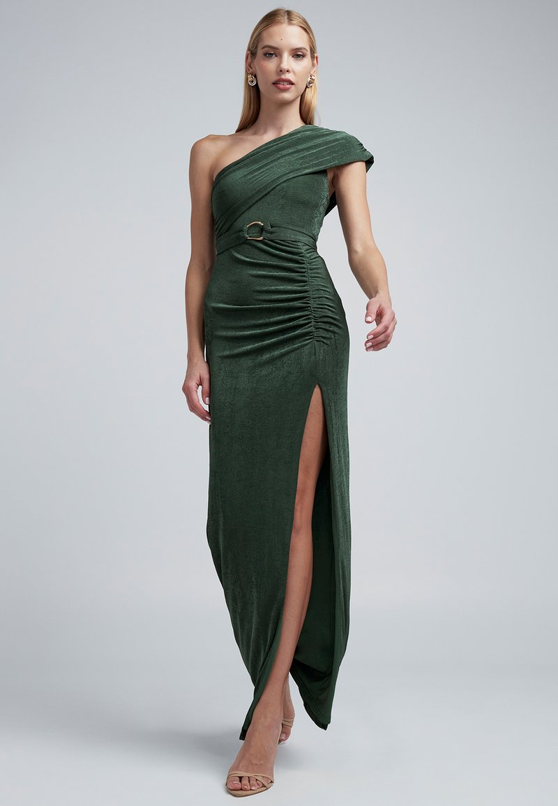 Jednokrčné zelené šaty z hodvábneho velúru so širokým rázporkom na boku, zdobené záhybmi a priliehavým strihom, doplnené dekoratívnym opaskom.